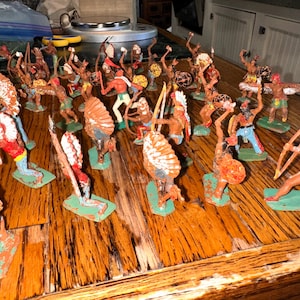 May include: A collection of hand-painted plastic toy figurines depicting Native American warriors in various action poses. The figures feature detailed headdresses, bows, spears, and shields, each mounted on a small green base, arranged in a group.