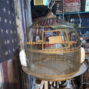 May include: A vintage brass birdcage with a round top and a wire mesh design. The cage has a wooden base and a brass label on the front. The cage is hanging from a chain and is surrounded by white ceramic ornaments.