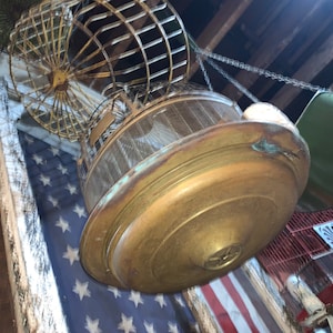 May include: A vintage brass birdcage with a wire mesh bottom and a round top. The cage is hanging from a chain and is suspended over a red, white, and blue American flag.