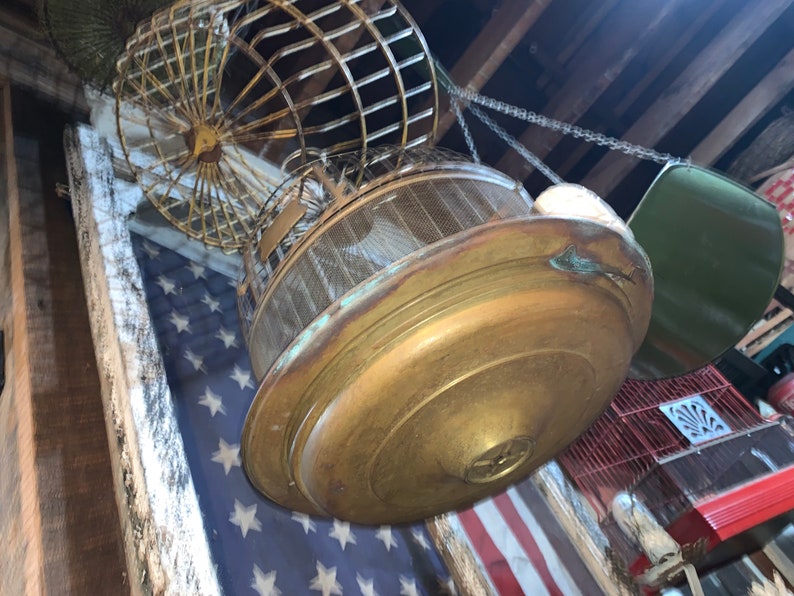 May include: A vintage brass birdcage with a wire mesh bottom and a round top. The cage is hanging from a chain and is suspended over a red, white, and blue American flag.