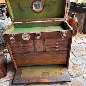 May include: An antique wooden tool chest with a hinged lid and multiple drawers. The interior is lined with green felt, and a small mirror is mounted inside the lid. The chest has metal latches and a decorative emblem on the front.