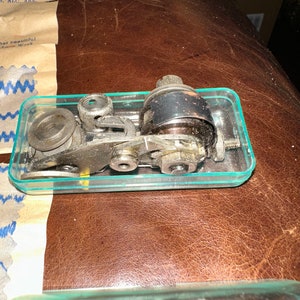 May include: A close-up of a vintage sewing machine part in a clear plastic container. The part is made of metal and has a silver and black finish. It has a small gear and a round metal piece with a hole in the center.