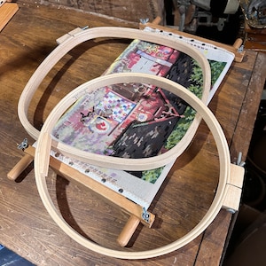 May include: Two wooden embroidery hoops, one inside the other, are mounted on a wooden frame. The hoops are used to stretch fabric taut for needlepoint or cross-stitch projects. The fabric is visible in the center of the hoops and features a colorful design.
