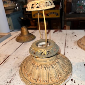 May include: A vintage metal lamp fixture featuring a wide, fluted base with a weathered gold finish. It includes a central socket assembly topped by a small, decorative, perforated dome cap supported by two thin, curved metal rods.