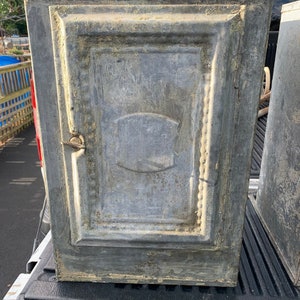 May include: A pair of antique, weathered, gray metal cabinets with ornate detailing. The cabinets have a single door and are closed. The cabinets are sitting in the back of a pickup truck.