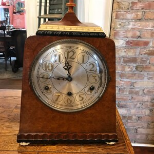May include: A vintage wooden mantel clock with a cream-colored face and black Roman numeral hour markers. The clock has a gold-colored trim and a wooden base with a decorative edge.