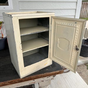 May include: A vintage cream-colored metal cabinet with a single hinged door that is open. The interior features two fixed shelves and a plain back panel. The exterior door has a decorative embossed rectangular frame and a small metal latch.