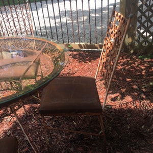 May include: A set of four rusty metal chairs and a glass-top table with a decorative metal frame. The chairs and table are sitting on a bed of red wood chips.