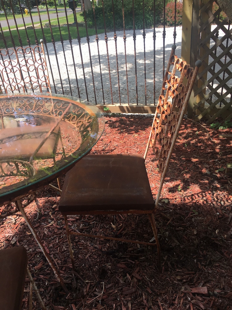 May include: A set of four rusty metal chairs and a glass-top table with a decorative metal frame. The chairs and table are sitting on a bed of red wood chips.