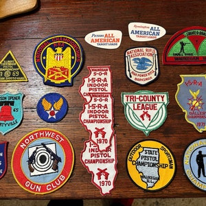 May include: A collection of 13 colorful embroidered patches featuring various shooting and gun club themes. The patches are mostly red, blue, yellow, and green with white lettering. Some of the patches feature the words "National Indoor NRA Championships", "Tri-County League", "State Pistol Championship", and "Northwest Gun Club".