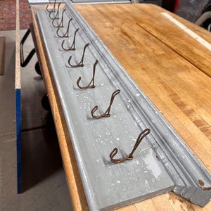 May include: A gray wooden shelf with a decorative trim and eight metal hooks. The shelf is painted a light gray color and has a distressed finish. The hooks are made of a copper-colored metal and are attached to the shelf with screws.