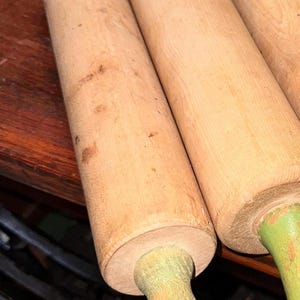 K&ouml;nnte beinhalten: Eine Sammlung von Nudelrollen aus Holz mit hellbraunen Walzen und gr&uuml;nen Griffen. Die Nudelrollen sind auf einer dunklen Holzoberfl&auml;che angeordnet und zeigen ihre nat&uuml;rliche Holzmaserung und ihren Vintage-Look.