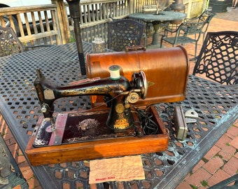 May include: Antique black and gold sewing machine with a wooden case. The machine is sitting on a black metal table with a wooden top. The machine has a spool of thread on the top and a needle in the front.