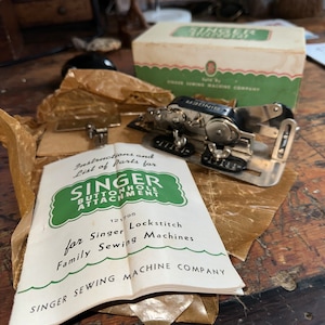 May include: A vintage Singer buttonhole attachment for Singer Lockstitch family sewing machines. The attachment is silver with black accents and comes with instructions and a list of parts. The packaging is brown paper with a green label.