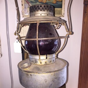 May include: An antique oil lamp with a red glass globe and a metal cage. The lamp has a hanging hook and a metal base.