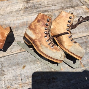 May include: A pair of vintage brown leather ice skates with laces and metal blades. The skates are worn and have a distressed look.