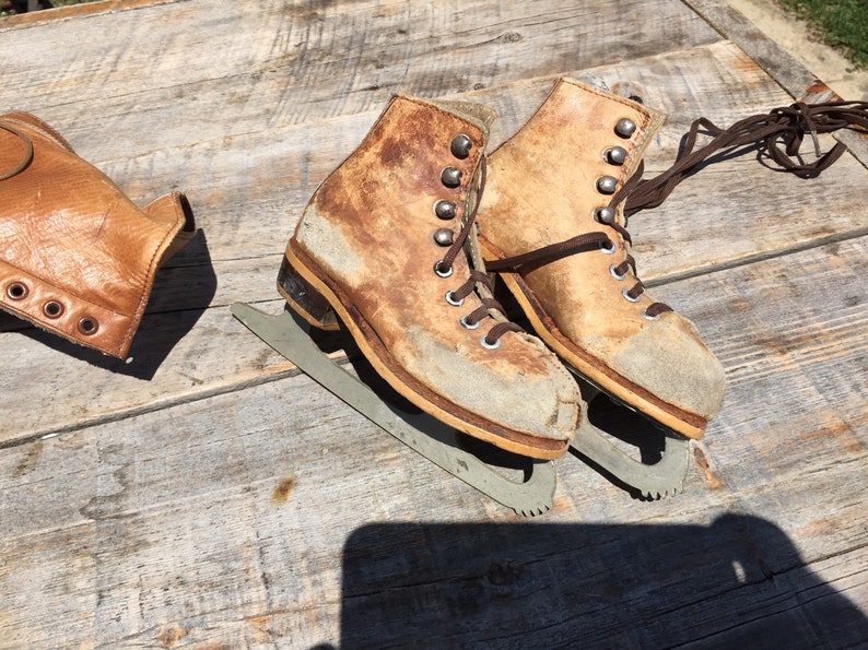May include: A pair of vintage brown leather ice skates with laces and metal blades. The skates are worn and have a distressed look.