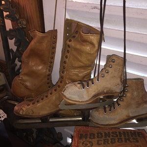 May include: A pair of vintage leather ice skates with brown leather boots and metal blades. The skates are hanging from a white wooden shutter.