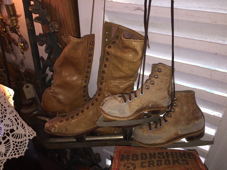May include: A pair of vintage leather ice skates with brown leather boots and metal blades. The skates are hanging from a white wooden shutter.