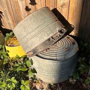 May include: Two round galvanized metal wash tubs with ribbed sides and metal ring handles. One tub is positioned at an angle resting on top of the other, which features a circular ridged pattern on its lid. The metal surfaces show signs of weathering.