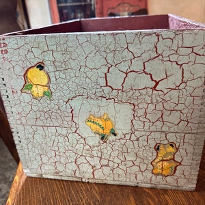 May include: A vintage wooden box with a distressed, light blue paint finish and red crackle glaze. The box features hand-painted yellow birds and a green frog.