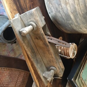 May include: A weathered, wooden tool with a rectangular body, metal hardware, and a handle. The tool is positioned on a dark wooden surface, with a partially visible wooden barrel in the background. The tool appears to be an antique woodworking or crafting instrument.