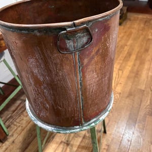May include: A large, antique copper bucket with a handle, showing signs of age and wear. The bucket is cylindrical and sits on a small, round, green metal stool. The copper has a rich, reddish-brown hue with areas of patina.