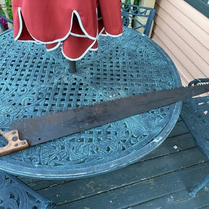 May include: A vintage hand saw with a wooden handle resting on a metal patio table with a decorative pattern. The saw is approximately 24 inches long.