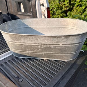 May include: An oval-shaped galvanized metal wash tub with a smooth, silver-grey finish. The basin features two horizontal embossed ridges running along the exterior walls, providing structural reinforcement to the wide, open-top container design.