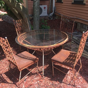 May include: A rusty metal patio set with a round glass-top table and four chairs. The chairs have intricate, ornate designs and are surrounded by red mulch.