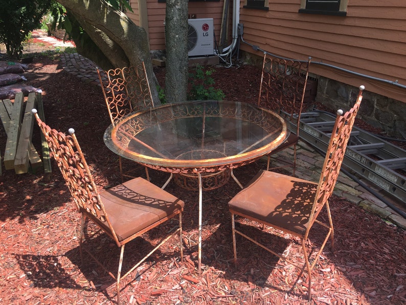 May include: A rusty metal patio set with a round glass-top table and four chairs. The chairs have intricate, ornate designs and are surrounded by red mulch.