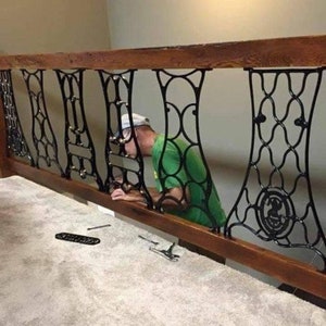 May include: A person is installing a black metal railing made from repurposed sewing machine parts. The railing is attached to a wooden beam and is being installed over a carpeted staircase.