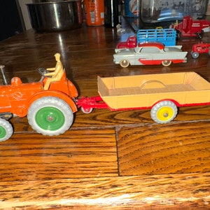 May include: A red toy tractor with a green steering wheel and a yellow figure behind the wheel, pulling a tan wagon with red wheels.