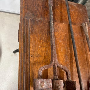 May include: A close-up of antique branding irons with wooden handles and rusty metal heads. The branding iron has three square heads. The background is a wooden surface.