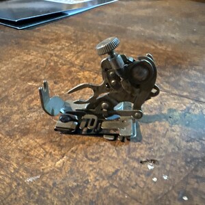 May include: A silver sewing machine foot with a small knob on top. The foot has a curved shape and several metal prongs.