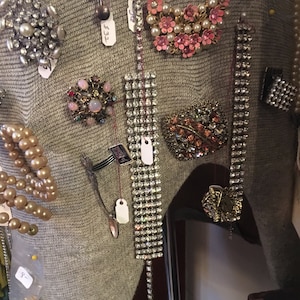 May include: A close-up of a gray sweater with various vintage brooches and jewelry pins. The pins are made of rhinestones, pearls, and other decorative elements. Some of the pins have price tags attached to them.