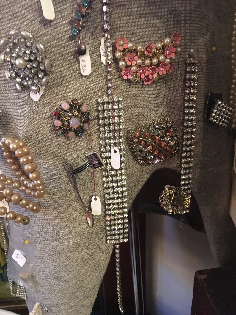 May include: A close-up of a gray sweater with various vintage brooches and jewelry pins. The pins are made of rhinestones, pearls, and other decorative elements. Some of the pins have price tags attached to them.