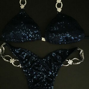 May include: A navy blue sequined bikini with crystal embellishments. The bikini top has a halter style with a chain-like design. The bikini bottom has a high-cut style with a chain-like design on the hips.
