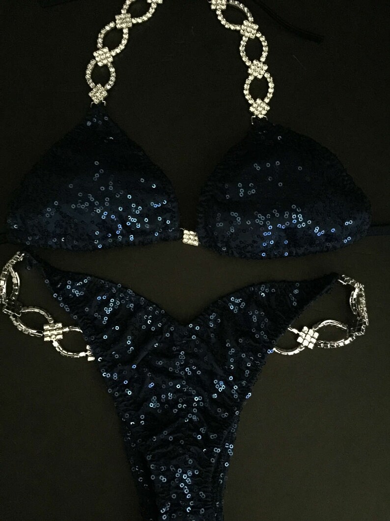 May include: A navy blue sequined bikini with crystal embellishments. The bikini top has a halter style with a chain-like design. The bikini bottom has a high-cut style with a chain-like design on the hips.