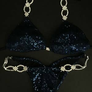 May include: A navy blue sequined bikini with crystal embellishments. The bikini top has a halter style with a chain-like design. The bikini bottom has a high-cut style with a similar chain-like design.
