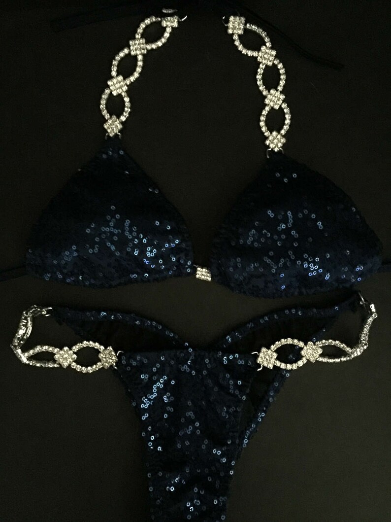 May include: A navy blue sequined bikini with crystal embellishments. The bikini top has a halter style with a chain-like design. The bikini bottom has a high-cut style with a similar chain-like design.