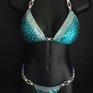 May include: Teal bikini set with rhinestone embellishments. The triangle top and the bottom are adorned with sparkling rhinestones. The straps and the waistline of the bottom feature a chain design with rhinestones. The bikini is displayed on a black mannequin.