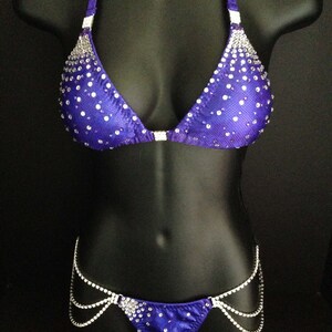 May include: A purple bikini with silver rhinestones and a silver chain detail. The bikini top has a triangle shape and the bottom is a high-cut style.