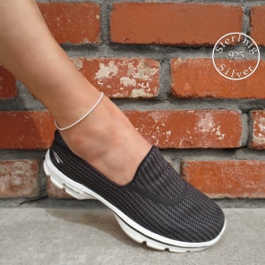 May include: A silver anklet with a delicate chain design. The anklet is worn on a person's ankle, with a black and white shoe visible in the background.