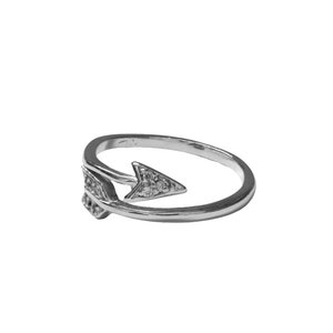 May include: A silver-colored ring with an arrow design. The arrow is adorned with small, clear gemstones. The ring's band curves around, creating an open design. The arrow points to the right.