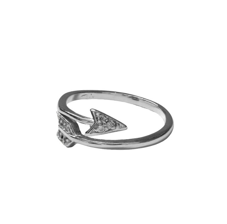 May include: A silver-colored ring with an arrow design. The arrow is adorned with small, clear gemstones. The ring's band curves around, creating an open design. The arrow points to the right.