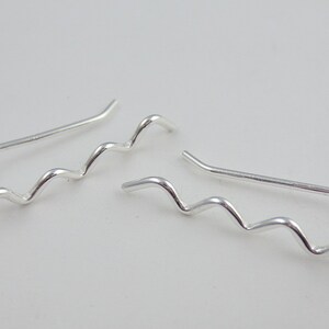 May include: A pair of silver-colored ear climbers with a wavy design. The earrings have a curved post that goes up the ear and a series of smooth, undulating curves. The jewelry is displayed against a plain white background.