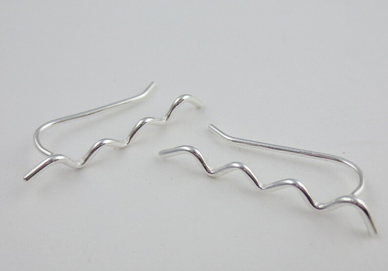 May include: A pair of silver-colored ear climbers with a wavy design. The earrings have a curved post that goes up the ear and a series of smooth, undulating curves. The jewelry is displayed against a plain white background.