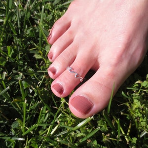 May include: A silver-colored toe ring with a wave design is worn on a foot with painted toenails. The foot is resting on a bed of green grass.