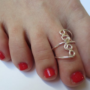 May include: A silver toe ring with a swirling design. The ring is worn on a foot with red nail polish.